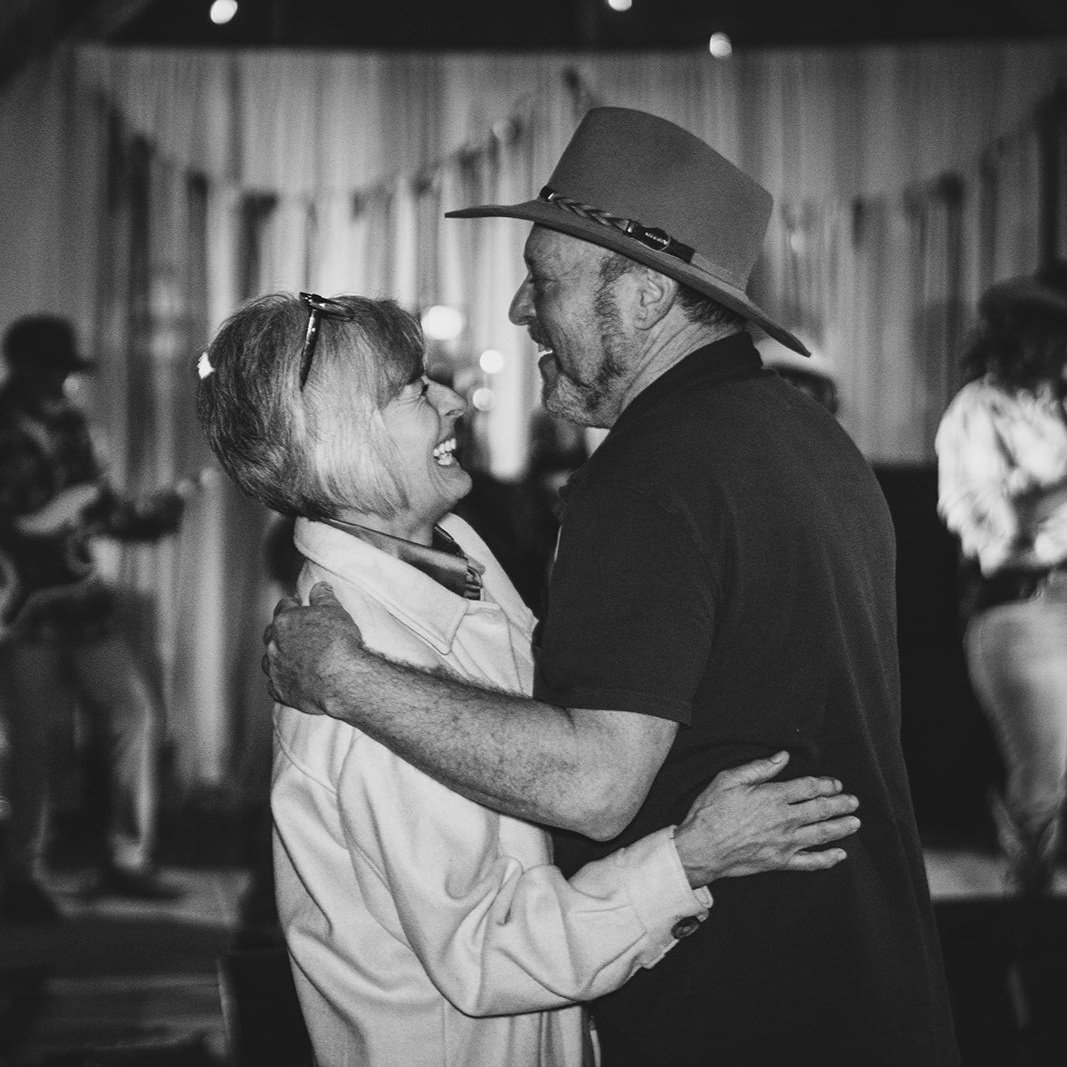 Couple dancing joyfully in a lively setting, showcasing happiness and connection, with soft lighting and blurred figures in the background.