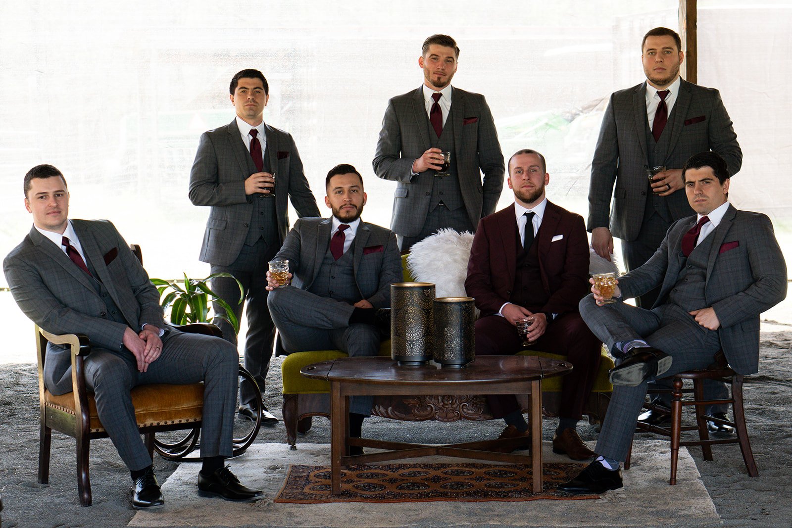 Group of eight men dressed in stylish suits, posing together indoors with drinks in hand. The setting features a vintage-style table and elegant decor, showcasing a sophisticated atmosphere suitable for a formal event or celebration.