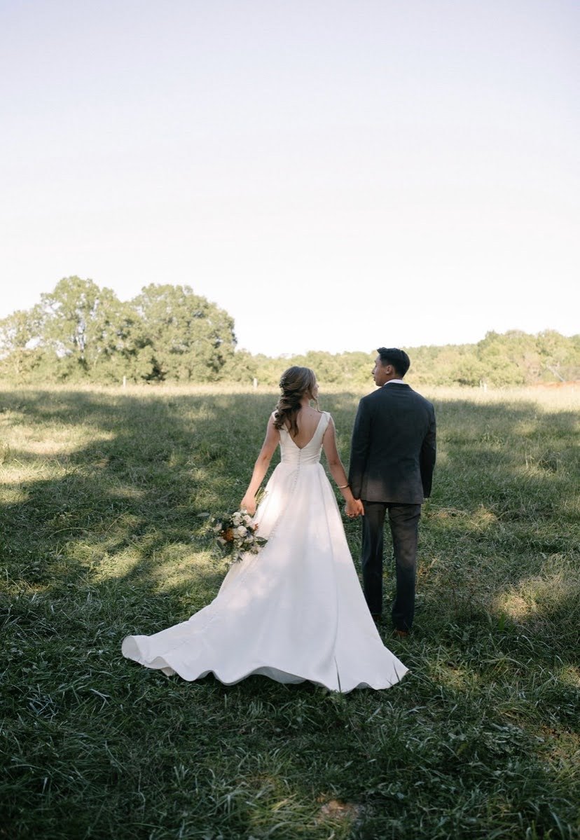Bride and groom holding hands, walking through a lush green field, with the bride wearing a flowing white wedding gown and holding a bouquet of flowers, captured in soft natural light.