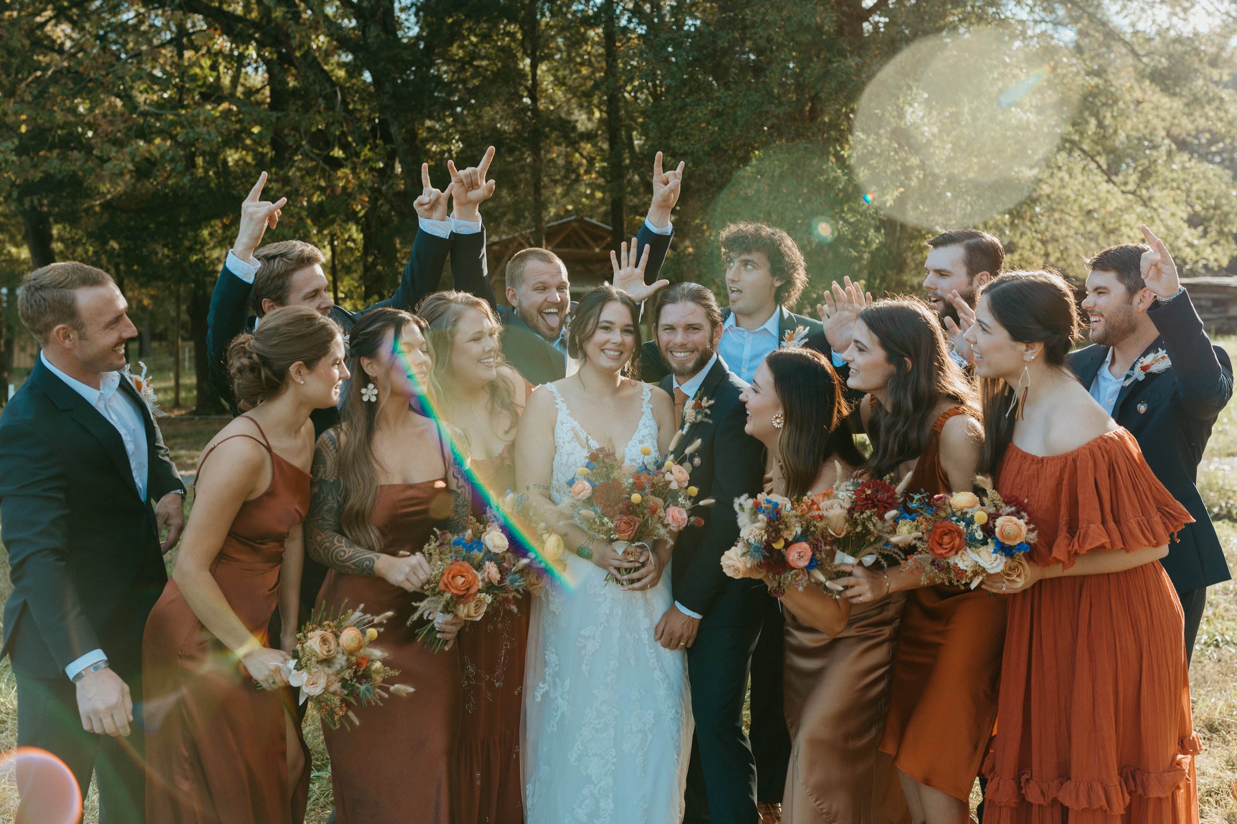 Group of joyful wedding guests celebrating outdoors, featuring the bride in a white gown surrounded by bridesmaids in earthy tones and groomsmen in suits, all holding colorful floral bouquets. The scene captures a lively and festive atmosphere with smiles and playful gestures in a natural setting.
