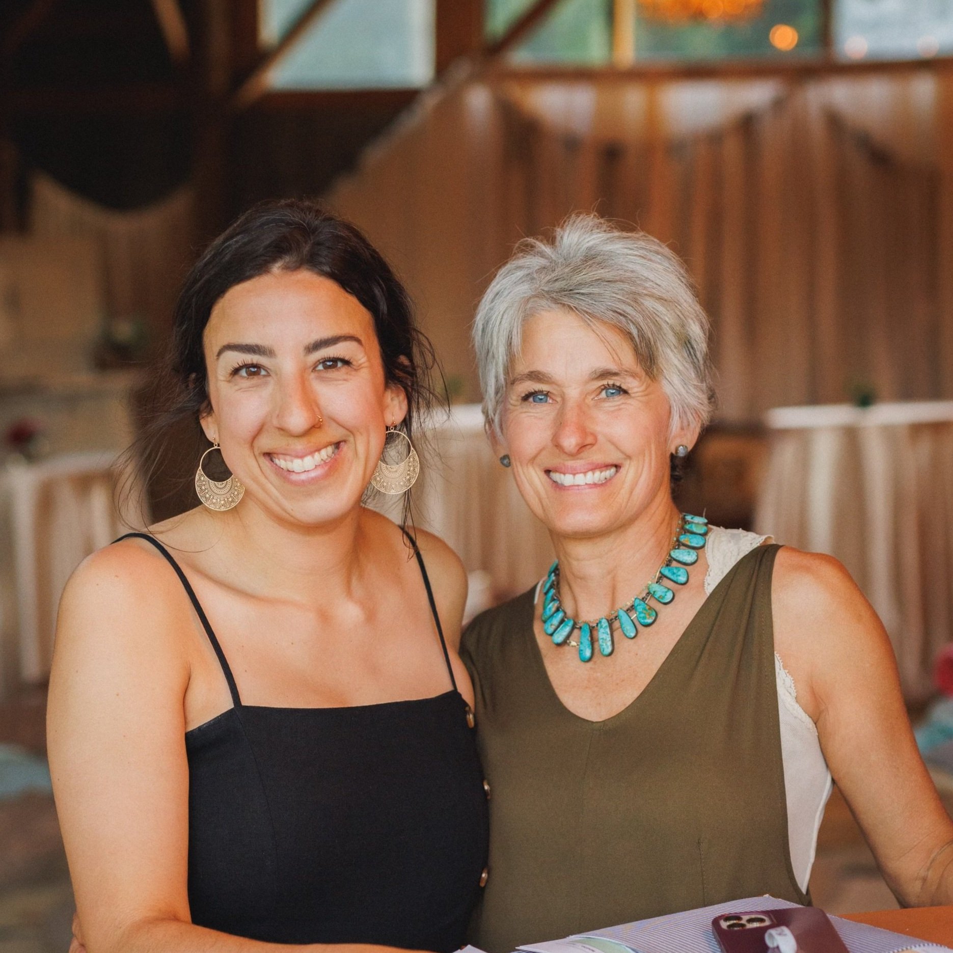 Two women smiling together in a rustic venue, showcasing a warm and joyful moment. The woman on the left wears a black sleeveless dress with large earrings, while the woman on the right has short gray hair and a turquoise necklace. The background features soft drapery and a hint of event decor, suggesting a celebration or gathering.