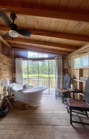 Cozy interior of a rustic cabin featuring a freestanding bathtub, wooden ceiling beams, and large windows with scenic views. The space includes a comfortable seating area with wooden furniture, creating a relaxing atmosphere.