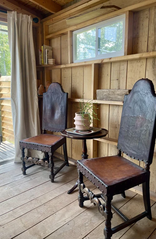 Cozy interior featuring two vintage wooden chairs with leather seats beside a small round table, adorned with a potted plant. Natural light filters through a window, highlighting the rustic wooden walls and warm ambiance. Ideal for a serene reading nook or relaxation space.