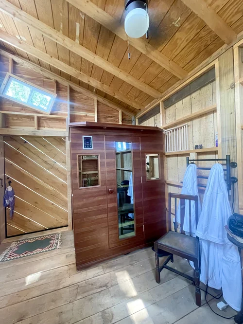 Interior of a cozy wooden cabin featuring a modern sauna, a wooden chair, and hanging robes, with natural light streaming through skylights and a decorative door mat visible.