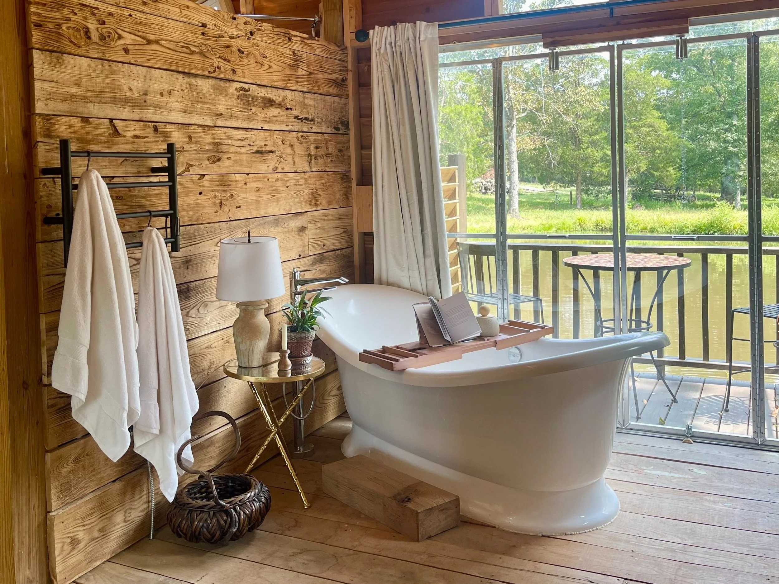 Modern bathroom interior featuring a freestanding white bathtub, wooden accent walls, and large windows overlooking a serene outdoor view. The space includes neatly hung white towels, a stylish lamp on a small golden table, and a decorative plant, creating a relaxing and inviting atmosphere.