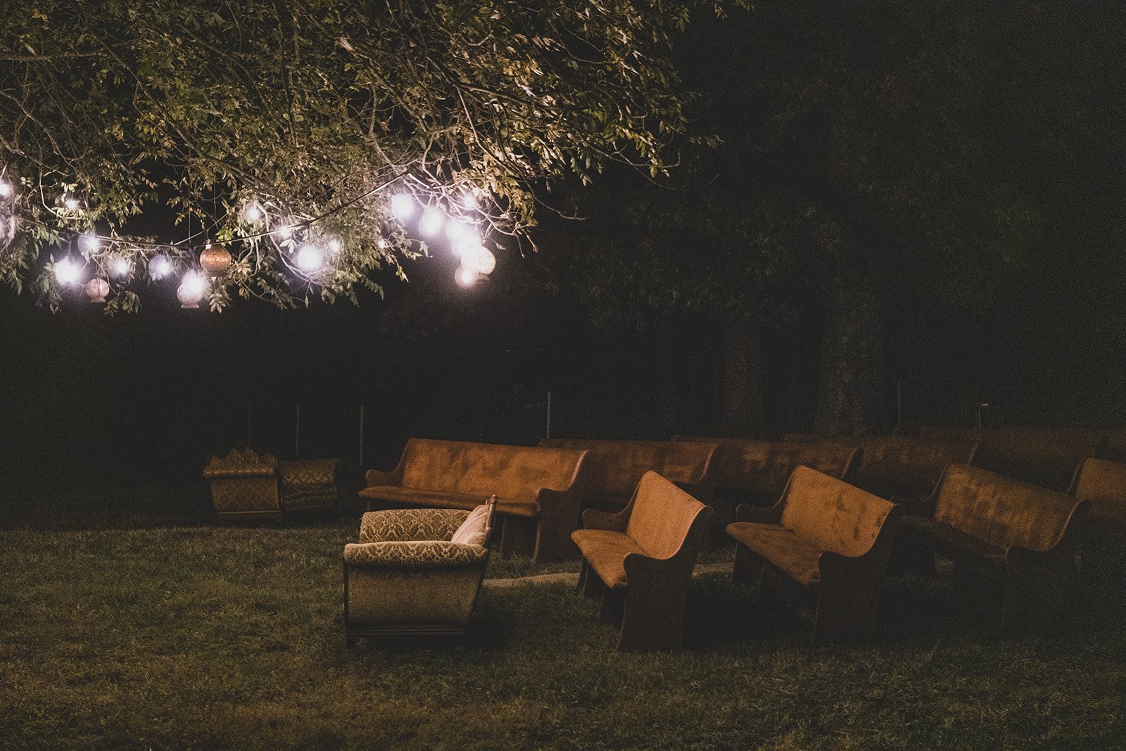 Cozy outdoor seating area illuminated by string lights, featuring vintage wooden benches and armchairs on a grassy field at night.