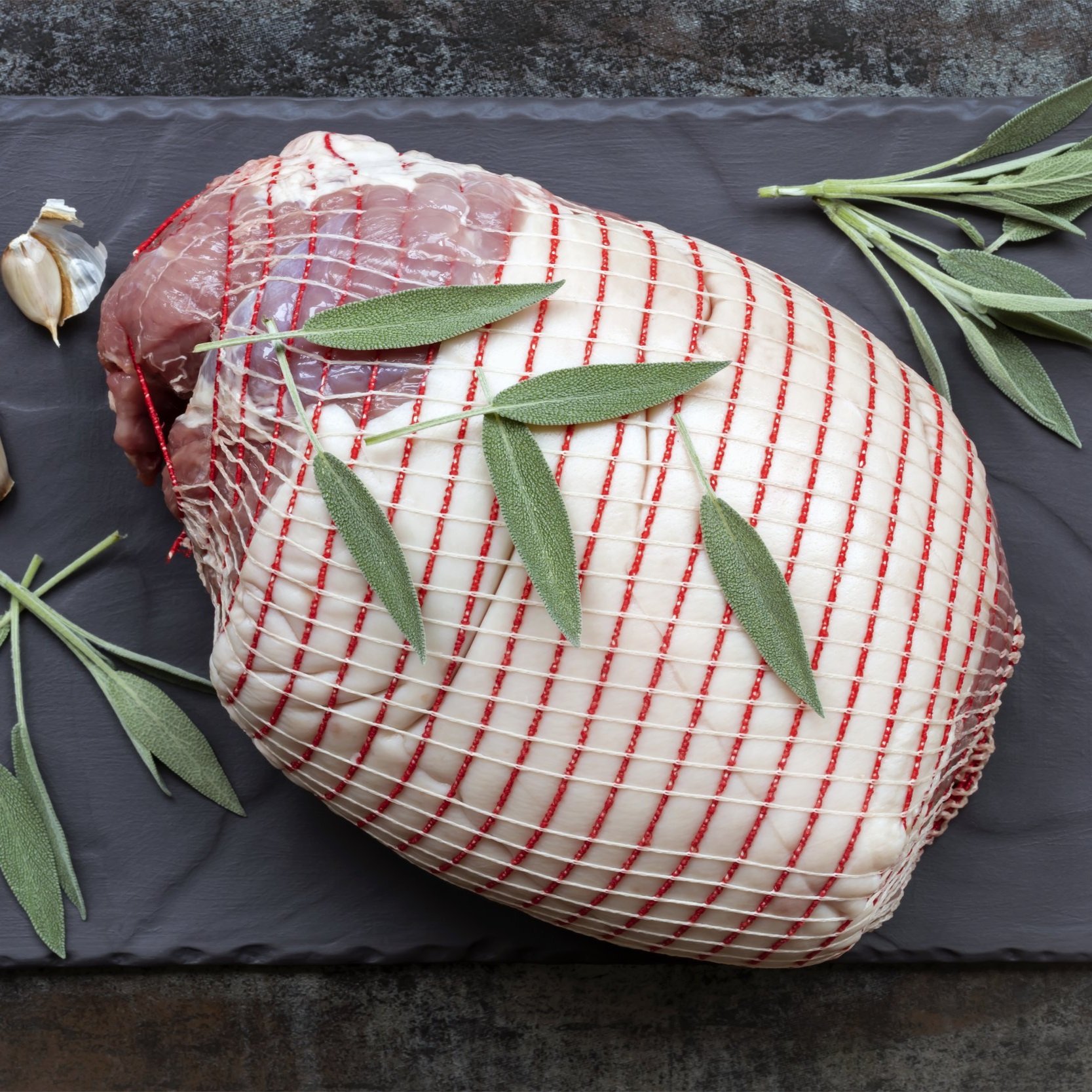 Raw meat joint tied with red string, garnished with fresh sage leaves and garlic cloves, displayed on a dark slate surface, ideal for holiday roasting or gourmet cooking.